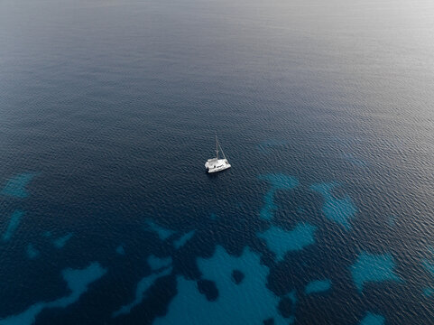 Aerial view of a white catamaran boat anchored in the crystal clear turquoise and deep blue waters of Cala rossa Favignana, Sicilia, Italy.