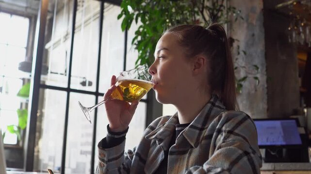 young woman sits in restaurant and drinks beer from glass and smiles, cute girl drinks alcohol in cafe