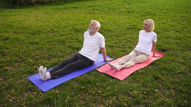couple of elderly seniors stretch and warm up on yoga mats in the park, gray-haired old pensioners exercise and do Pilates outdoors