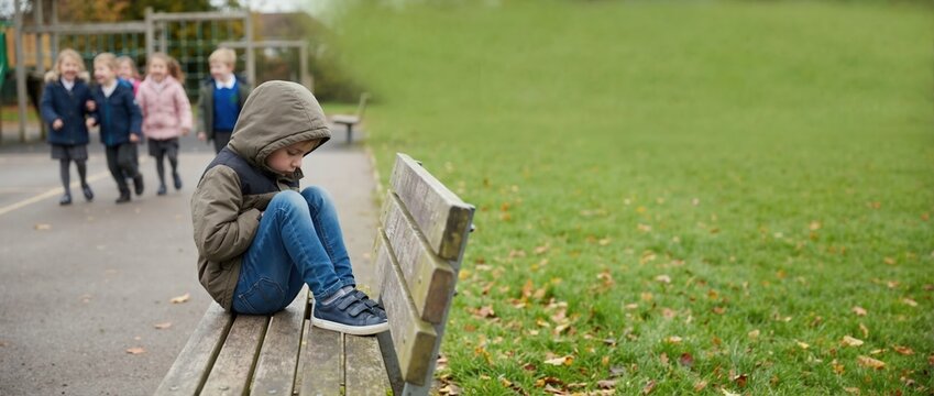 Sad boy sitting alone on bench while other children play in background. Lonely child excluded from group at playground. Concept of bullying and social isolation