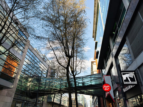 Modern glass skybridge and urban architecture in Manchester, UK with retail storefronts and reflective office buildings on a clear autumn day