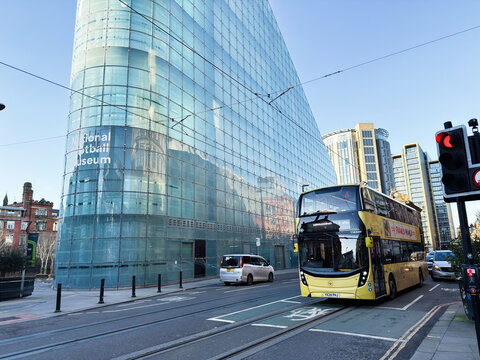 Yellow double decker bus passing the modern glass National Football Museum in Manchester, UK city street with pedestrians and tram tracks