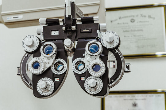 Manual phoropter refractor in professional ophthalmologist office setting. Wide shot of a full phoropter device against a clinic wall with blurred professional certificates in the background.