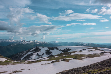 View from sunlit snowfield on hill top to snow cornice on rocky ridge against big snowbound mountain range far away under dramatic cloudy sky. Sunlight on snowy field in high mountains under clouds. © Daniil