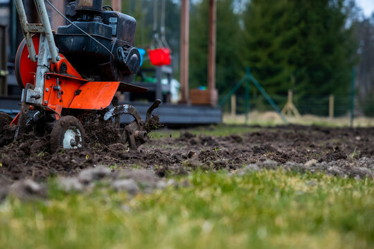 A bright orange rotary tiller churns wet soil, flinging clods near a deck and children's swings. Low angle shows wheels, belts, blades in soft cool light, early spring.