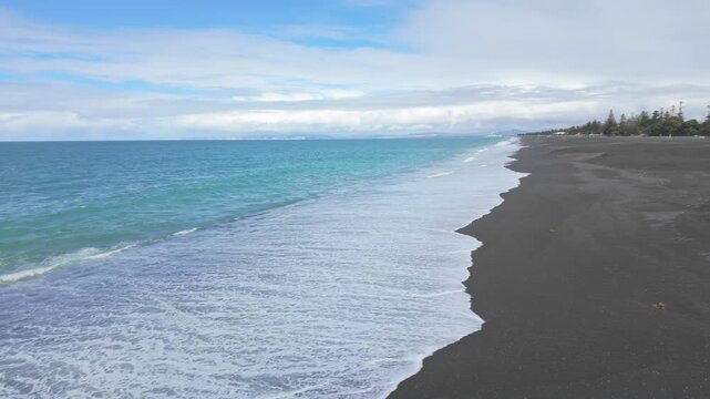A scenic view of the black sand beach in Napier, New Zealand, with the ocean waves gently lapping the shore and seagulls flying overhead. Marine Parade.