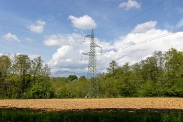 Obraz premium High voltage electricity pylon in spring rural landscape with ploughed field woodland and mountain background