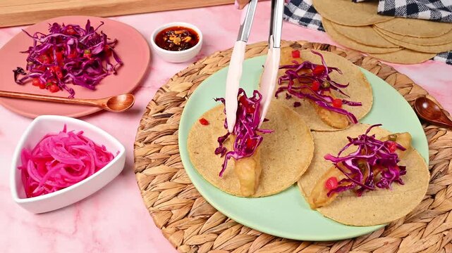 Hands preparing an Ensenada-style fish taco with a garnish of purple cabbage, tomato, and serrano peppers