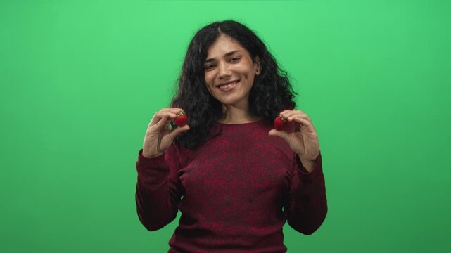 Young arab brunette woman holding strawberries between fingers and puckering lips in green studio; playful flirtation.