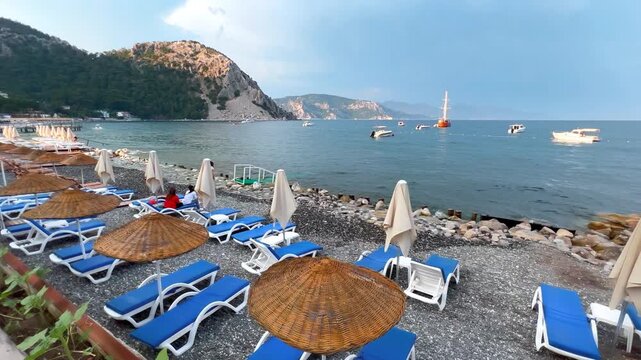 Beach resort with blue sunbeds and straw umbrellas on pebble shore in Turunc village, Marmaris, Turkey