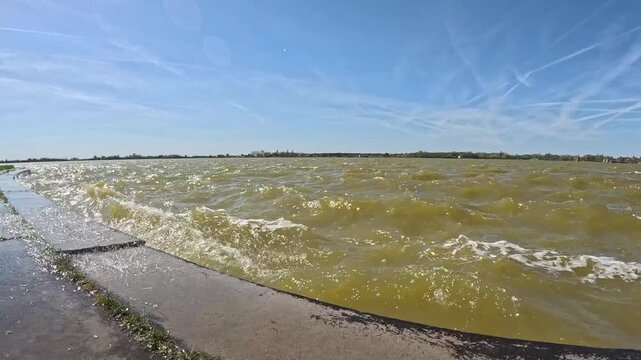 A detailed close-up shot focusing on the agitated surface of a large lake during a windstorm. The murky, yellowish water, filled with stirred-up sand, creates restless waves topped with thick white fo