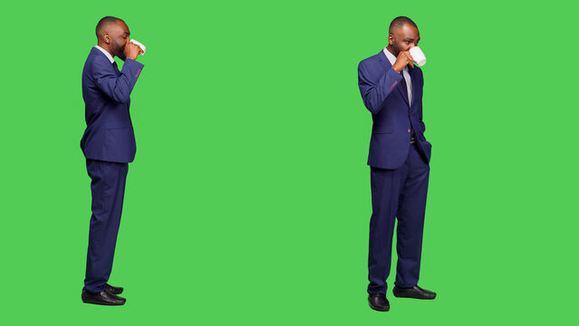 Company employee drinking coffee cup in studio, serving caffeine refreshment over full body greenscreen backdrop. Young manager enjoying refreshment drink, wearing formal suit on camera.