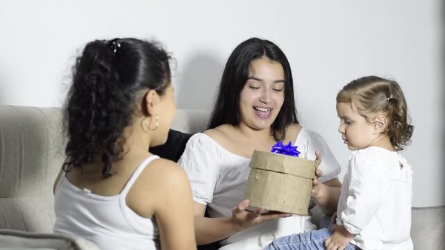 Little daughter gives a gift box to her Latina mother on the sofa sweet surprise and smiles at home.