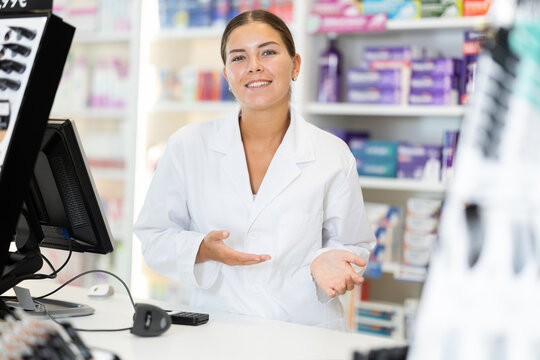Smiling young female apothecary standing in pharmacy store against background of shelves with medicaments