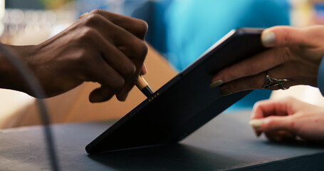 Fototapeta premium African american courier signing shipment document on tablet computer in modern boutique, discussing distribution details with worker. Manager working at online orders, preparing packages for delivery