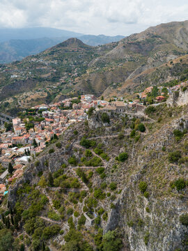Aerial view of the winding stone path on the steep cliffside overlooking the historic rooftops and rugged mountain landscape in Taormina, Sicily, Italy.