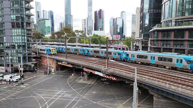 Drone footage of Melbourne Metro commuter train crossing elevated railway bridge through modern city center, surrounded by skyscrapers, streets and urban transport infrastructure in Australia.