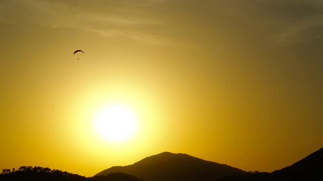 Paraglider flying towards the setting sun. Paraglider is enjoying a beautiful sunset over a mountain range. The sky is a vibrant yellow and orange, and the sun is a perfect circle