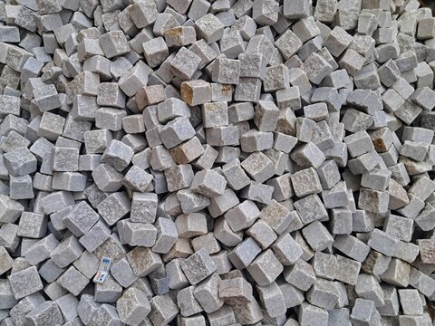 A top-down close-up view of a pile of gray granite paving stones, showing the natural texture and square shapes used for traditional European streets.