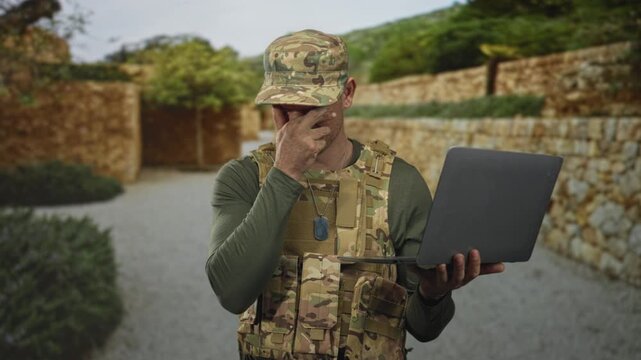 Man in camouflage uniform and cap holds laptop, crosses fingers and touches chin showing dog tags in building courtyard; hopeful.
