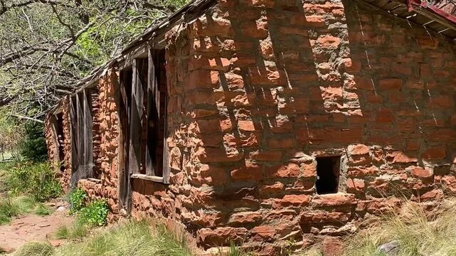 Cinematic view of a historic red stone cabin with a collapsed wooden roof. Surrounded by lush forest and majestic Sedona mountains under a blue sky. Sunny day, hiking trail, and ghost town vibe.