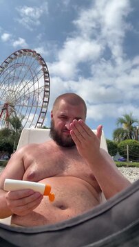 Vertical travel video. 60 fps. Mature tanned bald white fat naked man with beard applies moisturizer, milk balm, sunscreen cream to face on sun lounger on sea beach, ocean shore. Ferris wheel
