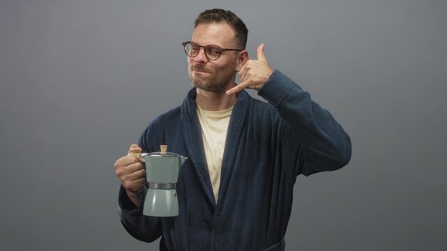 Man in robe holds a moka pot coffee maker and makes a thumb and pinky call gesture with his hand in studio; morning calm.