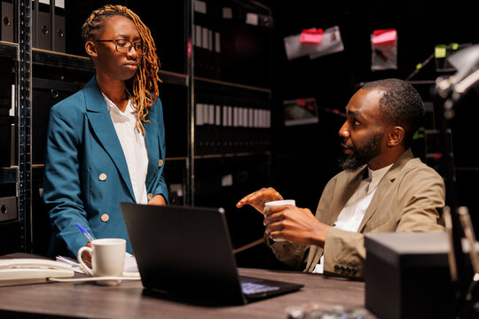 African american cops team working in archive to solve crime case. Man detective sitting at desk near shelf full of folders and files, discussing insight with woman partner at night time