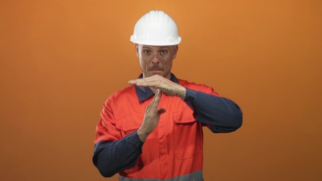Man in white hardhat and orange safety uniform making a timeout hand gesture with visible tattooed hands against orange backdrop in studio; alert safety.