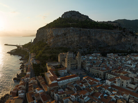 Aerial view of the historic Cefalu Cathedral and the massive limestone cliff of La Rocca overlooking the coastal town at sunset in Cefalu, Sicily, Italy.