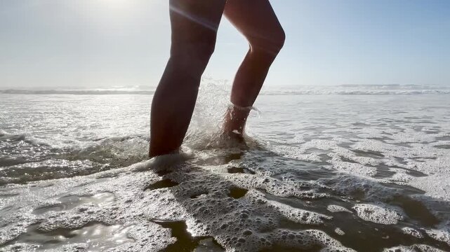 Close up of a woman walking barefoot in the Atlantic Ocean waves. Water splashes around her feet. Summer vacation, freedom, relaxation, nature, travel, and beach lifestyle