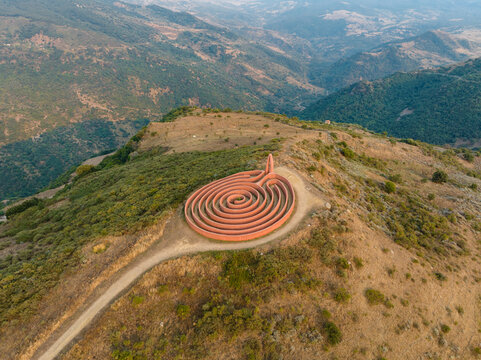 Aerial view of the Labirinto di Arianna sculpture on a hilltop surrounded by rolling green mountains and hazy valleys in Castel di Lucio, Sicily, Italy.