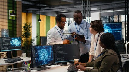 Naklejka na ściany i meble Multiethnic technicians use devices in AI startup office to train predictive models for language processing tasks. Multiracial coworkers use deep learning technical expertise for tasks, camera A