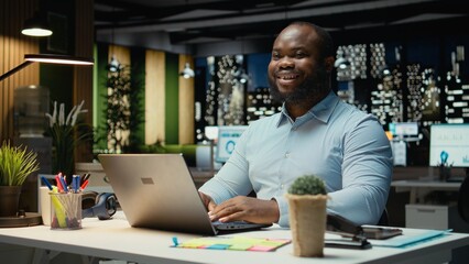Fototapeta premium Portrait of african american joyful guy manages emails and administrative tasks, documenting insights for a multinational company. Meeting business objectives late at night in office.