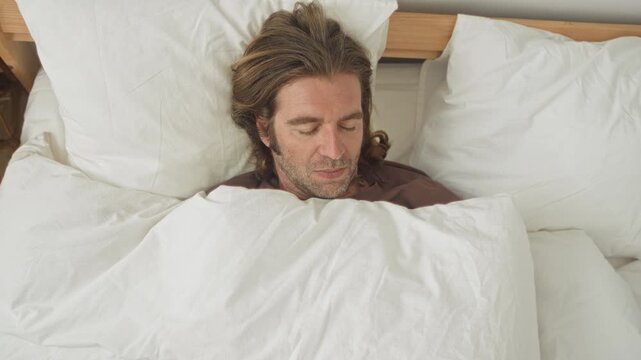 Man in bed clutching white duvet with hands, peeking over covers while looking down toward sheet in bedroom; unease reflection.