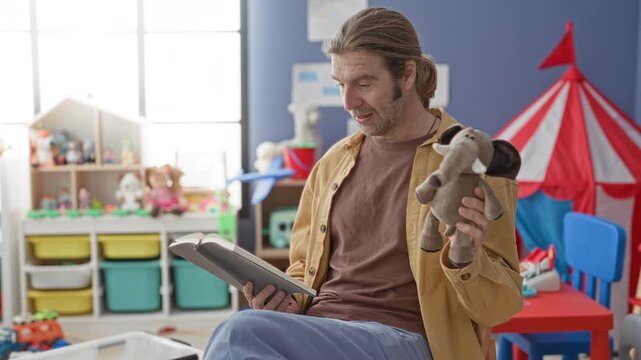 Man holds a stuffed elephant puppet and reads aloud from an open book while seated in a classroom play area with toys and storage bins; nurturing storytelling connection.