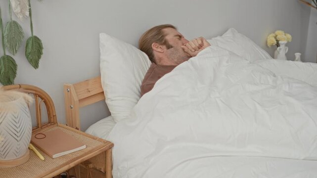 Man yawning, hand over mouth on bed in building, covered by white duvet and pillow; sleepiness morning.