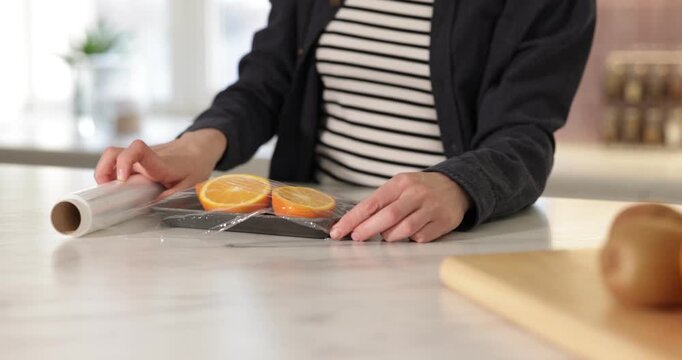 Woman wrapping container with fresh orange slices in cling film at light marble table, closeup. Camera moving