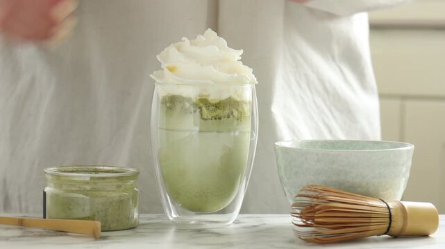 Woman adding whipped cream to drink and sifting matcha powder at white marble table indoors, closeup