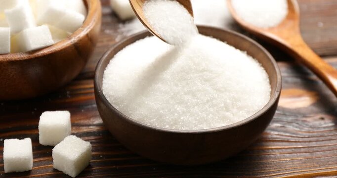 Pouring granulated sugar into bowl and cubes at wooden table, closeup