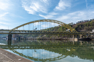Obraz premium Fort Pitt Bridge Crosses Monongahela River in Pittsburgh With Clear Sky and Green Hillside in View During Daylight Hours