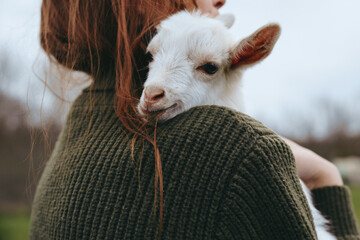 Affectionate woman in a chunky sweater carries a playful goat kid on her shoulder outdoors, capturing a wholesome countryside lifestyle moment, warm, gentle, and intimate. © SHOTPRIME STUDIO