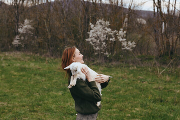 Obraz premium A woman outdoors in a green field holds a small goat kid close, smiling softly. Warm light and blooming trees frame this tender moment of countryside bonding and gentle animal care.