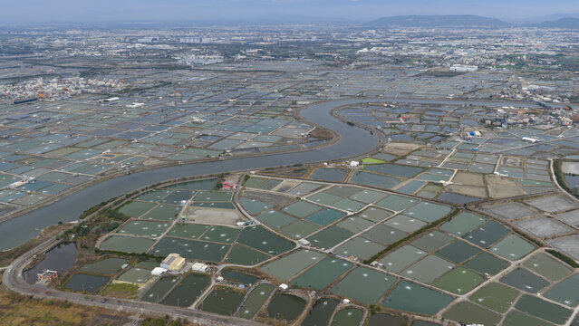 Aerial view of a vast network of rectangular fish ponds and aquaculture farms divided by narrow paths with a winding river under a hazy sky in Yonghua Village, Kaohsiung City, Taiwan.