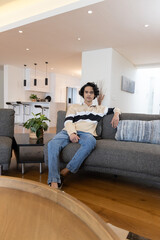 Obraz premium Asian adult man sitting on gray sofa in modern open-plan home with kitchen island and plant