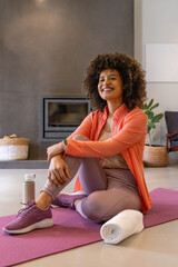 Naklejka premium African American woman in coral jacket sitting on purple mat at home, holding water bottle, towel