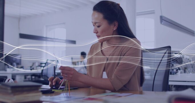 Working Korean designer in tan blouse using stylus on tablet at open office, holding color swatches