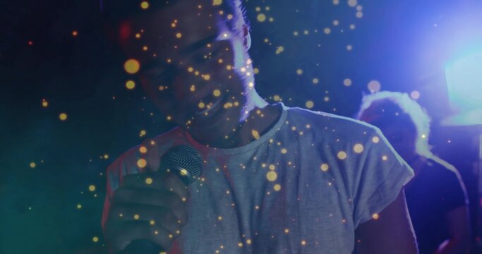 Singing female singer holding microphone on club stage, wearing light tee amid golden bokeh lights