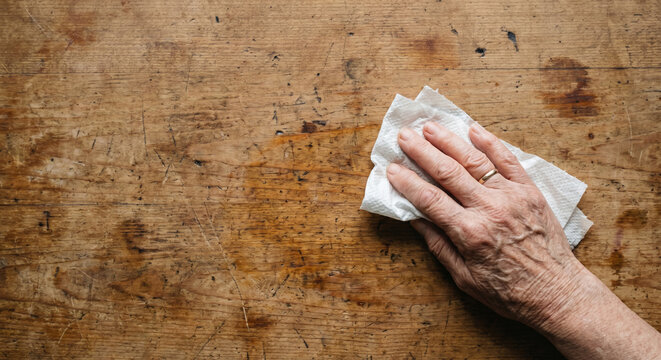 Close-up top view of a mature wrinkled hand wiping a dirty wooden table with a paper towel, household cleaning and hygiene concept.