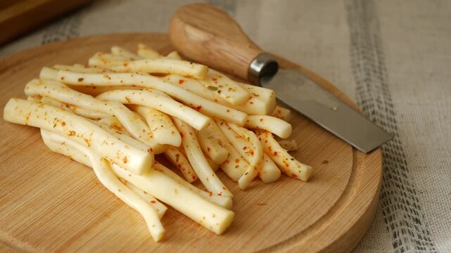 Spiced string cheese snacks on wooden board with metal knife rotating slowly in video clip.
